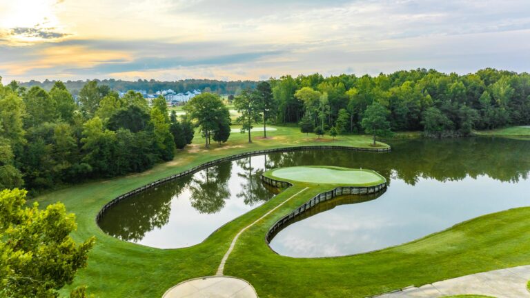 Aerial view of a golf course with a curved water hazard around the greens and trees, at sunset.