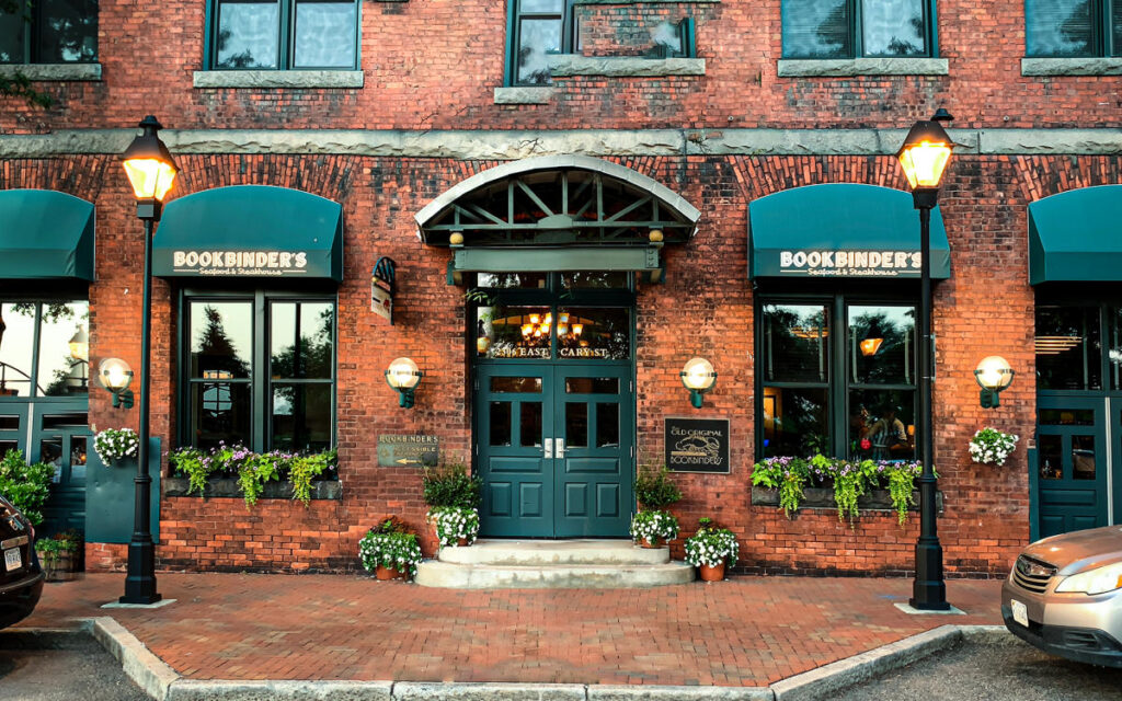 Brick storefront for Bookbinder's Seafood & Steakhouse with green awnings, lamps, and potted plants framing the entrance to a brick building.